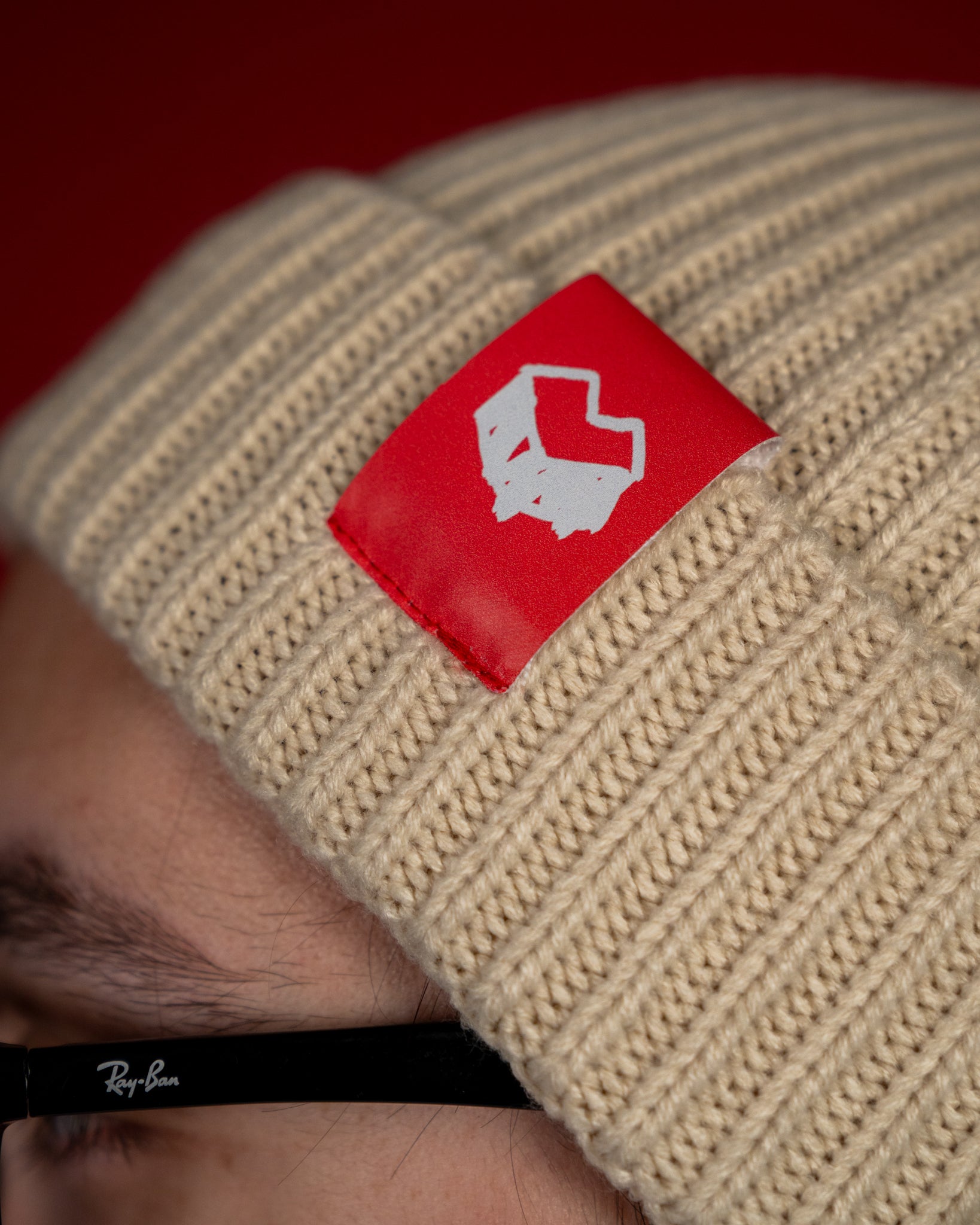 Nahaufnahme einer beigefarbenen Strickmütze mit breiter Rippstruktur. Auf der umgeschlagenen Krempe befindet sich ein rechteckiges, rotes Label mit weißem „Loot für die Welt“-Logo. Das weiche Strickmaterial wirkt warm und elastisch – ideal für kühle Tage.