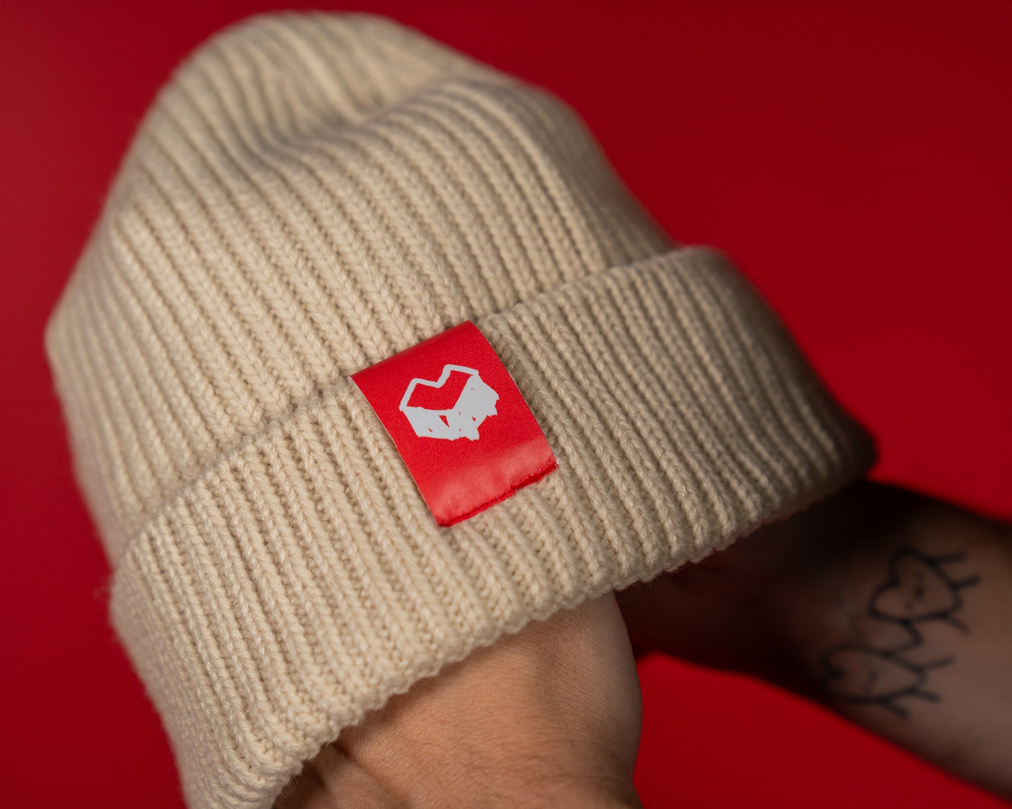 Beigefarbene Strickmütze mit breiter, umgeschlagener Krempe. Auf der Vorderseite befindet sich ein rotes Label mit weißem „Loot für die Welt“-Logo. Die grobe Rippstruktur sorgt für ein weiches, warmes Tragegefühl. Die Beanie kombiniert schlichte Streetwear-Optik mit einem markanten Farbakzent am Logo-Patch.