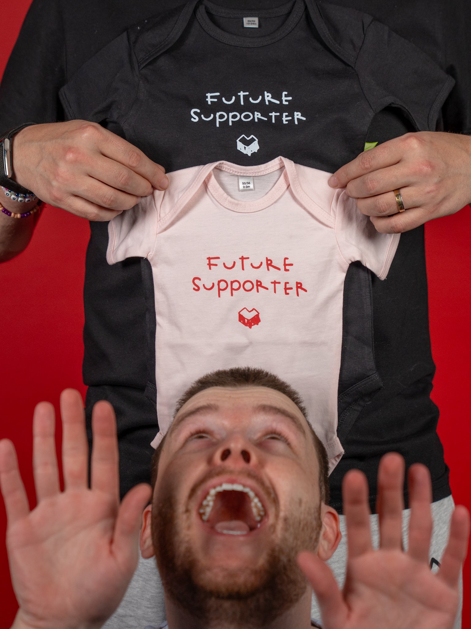 Zwei Babybodys aus weicher Baumwolle in den Farben Schwarz und Hellrosa. Beide Modelle zeigen den handschriftlich gestalteten Schriftzug „FUTURE SUPPORTER“ auf der Brust, ergänzt durch das „Loot für die Welt“-Logo darunter – in Weiß auf dem schwarzen und in Rot auf dem rosa Body. Beide Bodys besitzen kurze Ärmel, einen Rundhalsausschnitt und Druckknöpfe im Schritt für bequemes An- und Ausziehen. Das Bild zeigt die beiden Varianten gemeinsam, um die Farbunterschiede und das identische Design zu verdeutlichen