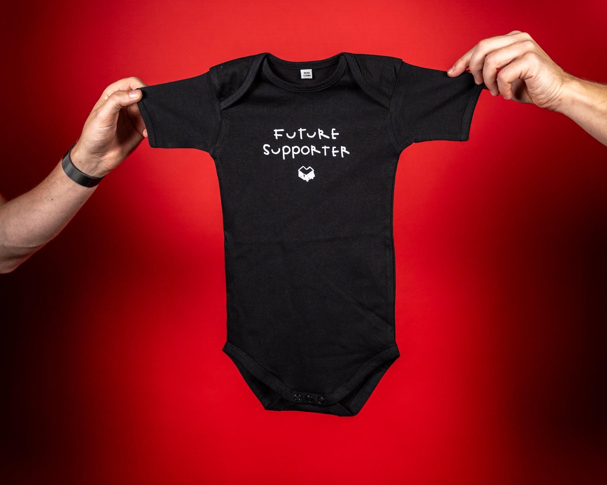 Schwarzer Babybody aus weicher Baumwolle mit kurzen Ärmeln und Rundhalsausschnitt. Auf der Vorderseite steht in weißer, handschriftlicher Schrift der Text „FUTURE SUPPORTER“, darunter das weiße „Loot für die Welt“-Logo. Der Body hat Druckknöpfe im Schrittbereich für einfaches An- und Ausziehen und ist angenehm elastisch für optimale Bewegungsfreiheit.