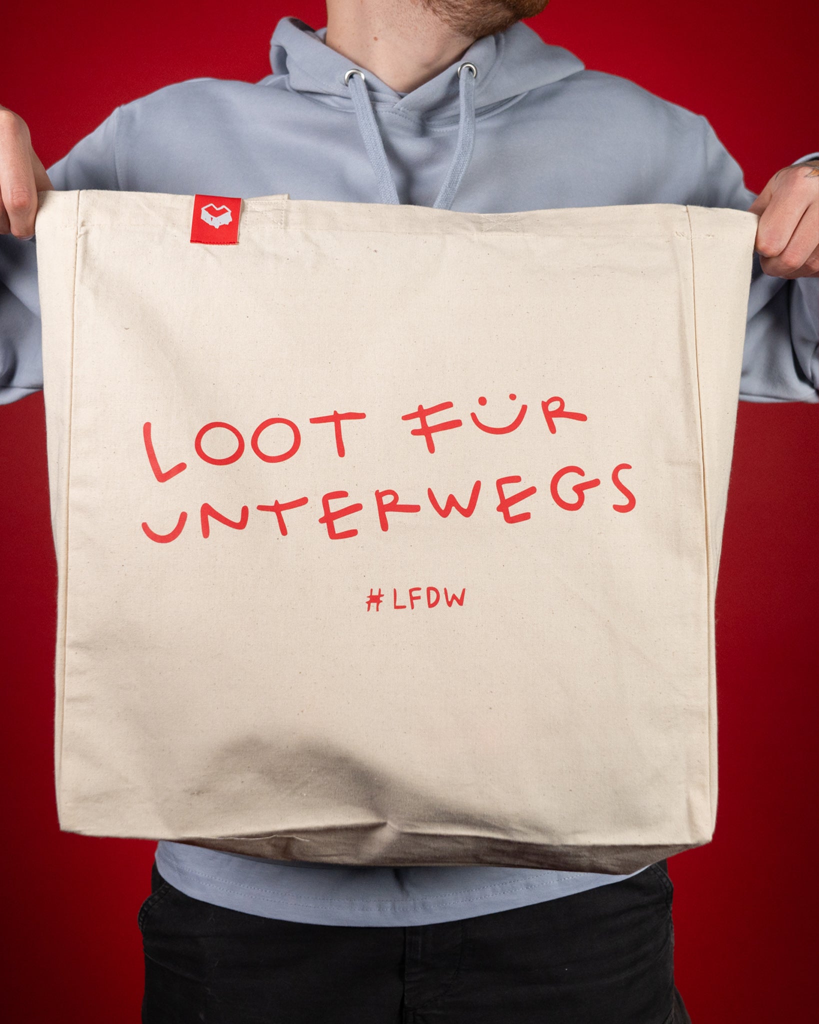 Beigefarbene Stofftasche aus robuster Baumwolle mit rotem Aufdruck „LOOT FÜR UNTERWEGS“ und dem Hashtag „#LFDW“ auf der Vorderseite. Oben an der Naht ist ein kleines rotes Label mit weißem Symbol angebracht. Die Tasche hat zwei lange Henkel und bietet viel Stauraum für Einkäufe oder Alltagsgegenstände.