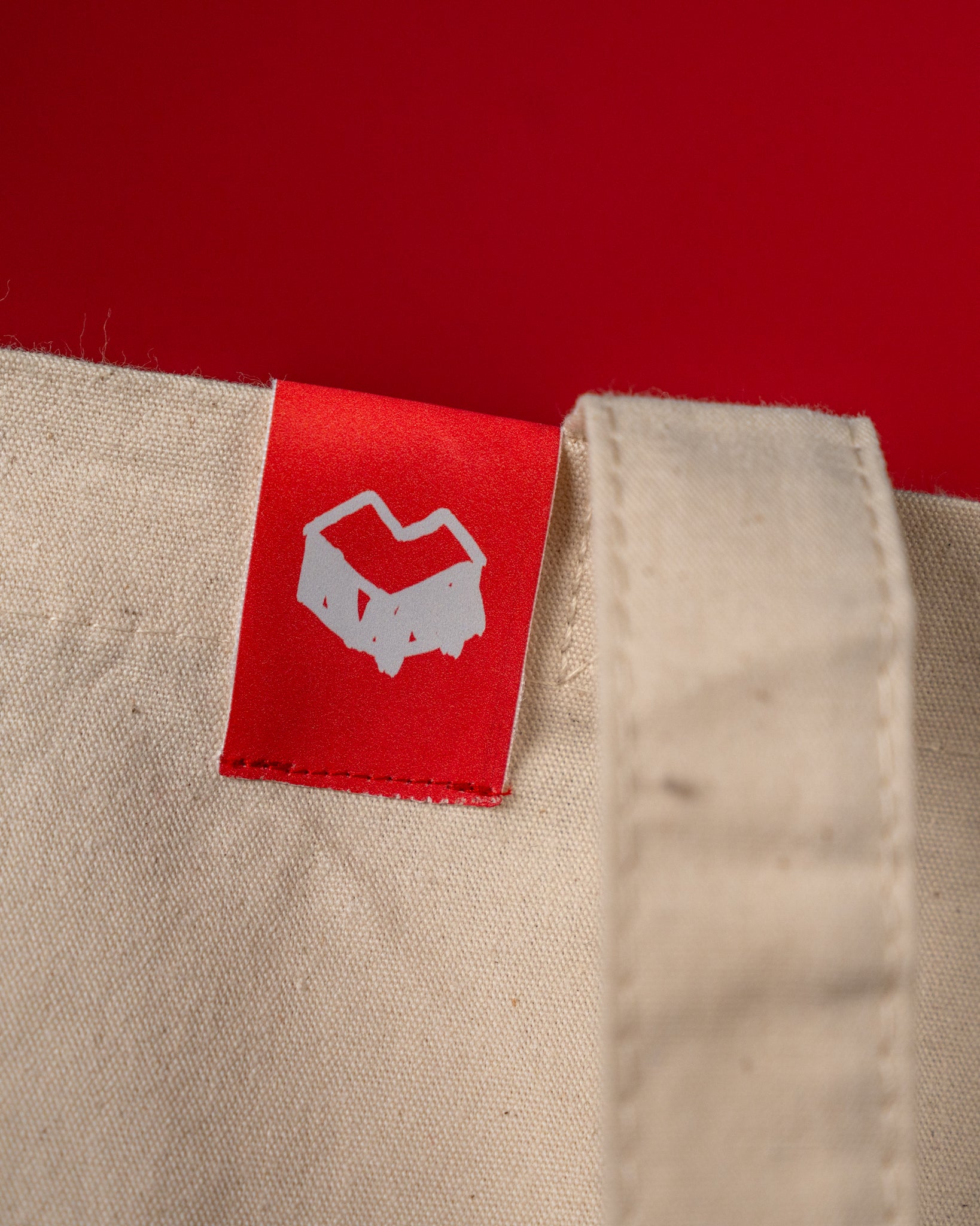 Detailaufnahme einer beigefarbenen Stofftasche mit fest vernähtem roten Label an der oberen Kante. Das Etikett zeigt ein weißes, grafisches Symbol auf rotem Hintergrund. Der Stoff besteht aus grob gewebter Baumwolle und wirkt stabil und hochwertig verarbeitet.