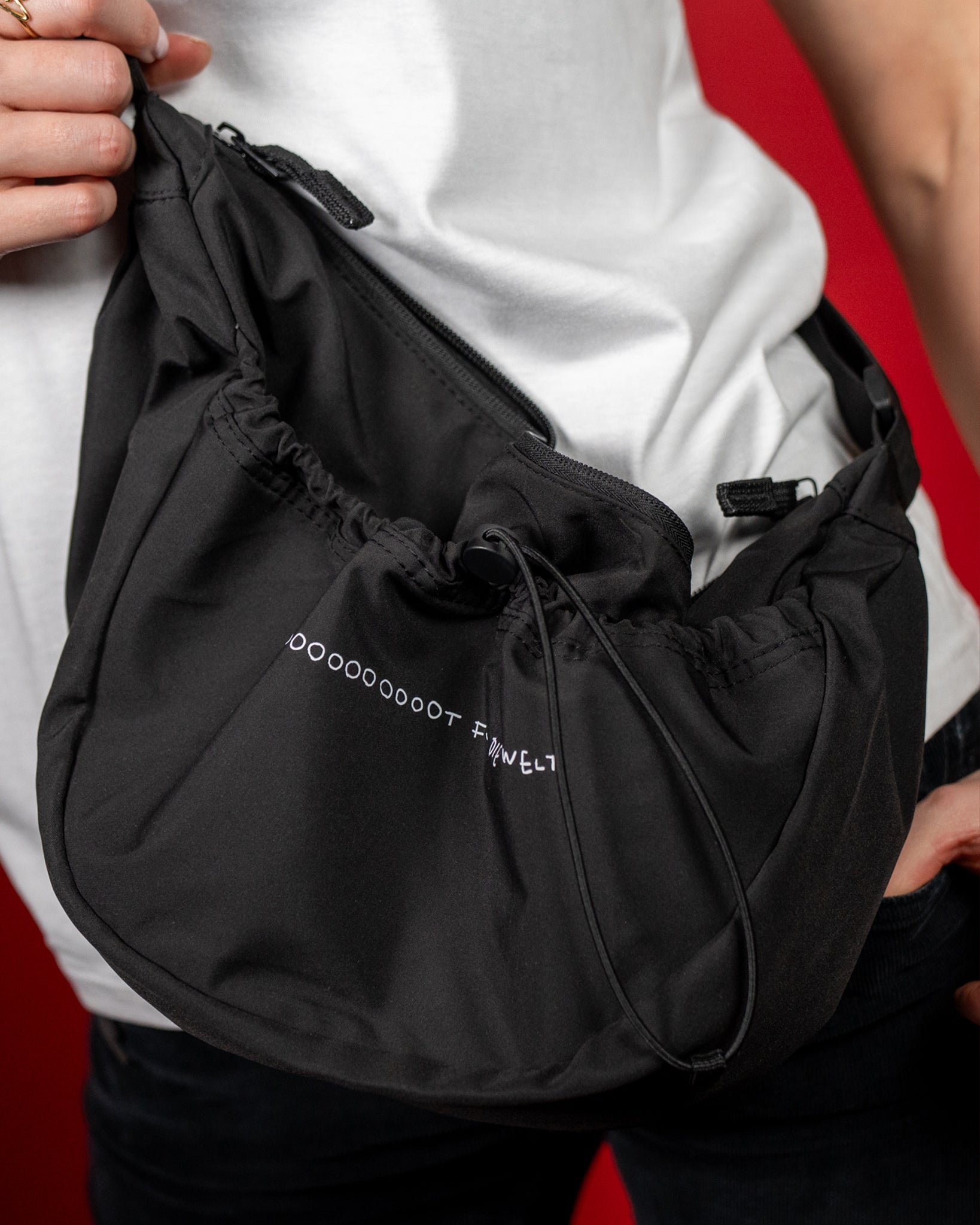 Schwarze Umhängetasche aus glattem, leicht strukturiertem Stoff mit verstellbarem Gurt. Die Tasche besitzt ein Hauptfach mit Reißverschluss sowie eine geraffte Fronttasche mit elastischem Kordelzug. Auf der Vorderseite ist in kleiner, weißer Schrift das Logo „LOOT FÜR DIE WELT“ aufgedruckt. Die weiche Form und das funktionale Design verleihen der Tasche einen modernen, sportlichen Look.