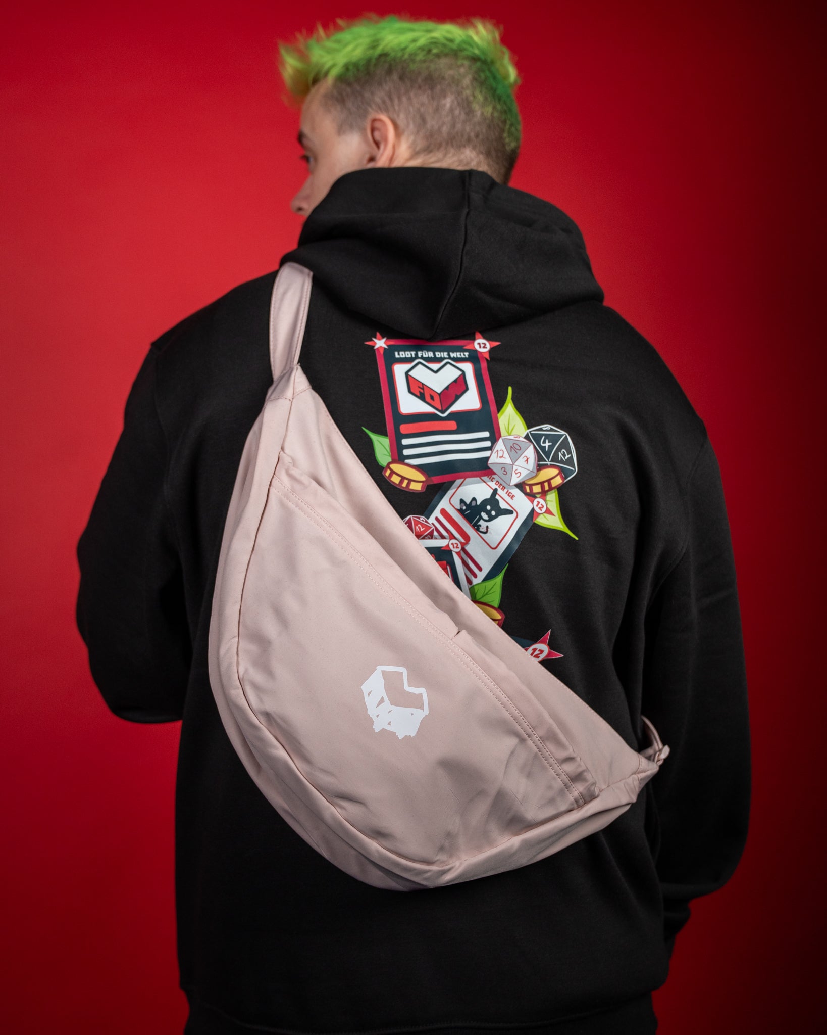 Hellrosa Umhängetasche mit weißem Logo-Print in Form eines stilisierten Würfels auf der Vorderseite. Die Tasche wird quer über dem Rücken getragen und besteht aus glattem, leichtem Stoff mit Reißverschlussfach. Das Design wirkt schlicht und modern, ideal für den Alltag oder Events. Im Hintergrund ist ein schwarzer Hoodie mit farbigem „Loot für die Welt“-Print zu sehen.