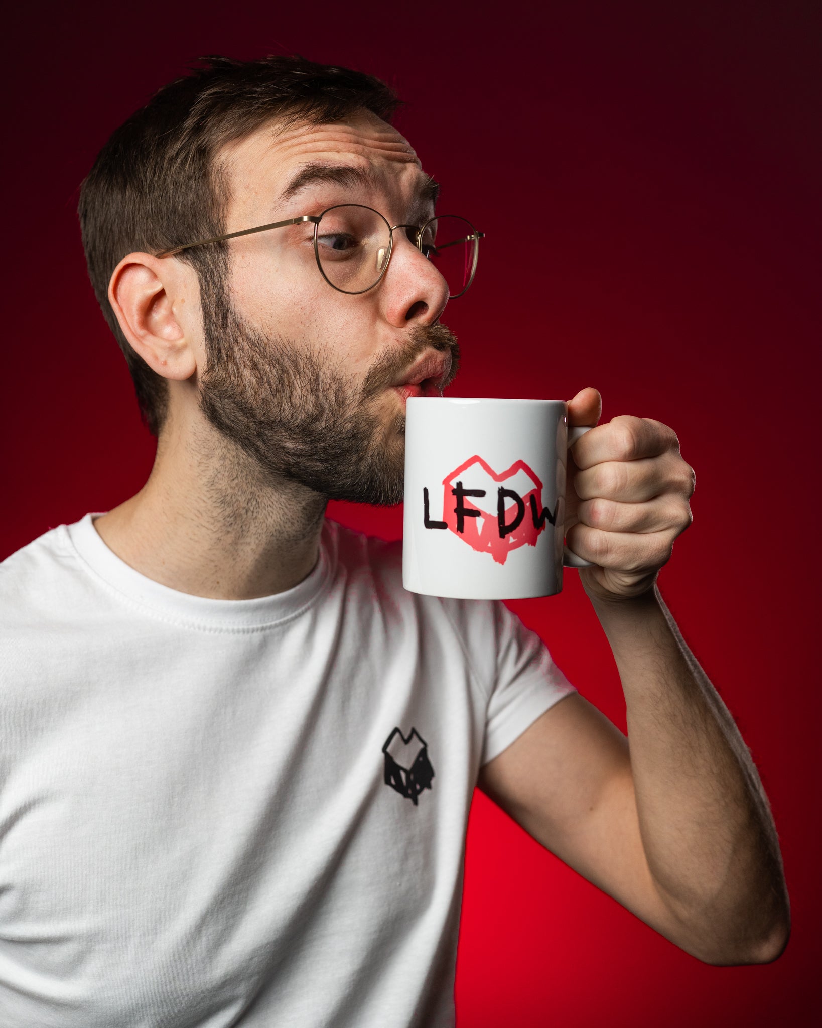 Weiße Keramiktasse mit schwarzem Schriftzug „LFDW“ auf einem roten, herzförmigen Farbklecks. Die Tasse wird seitlich gehalten, der Aufdruck ist klar sichtbar. Das Design steht für „Loot für die Welt“ und kombiniert verspielte Typografie mit einem markanten Symbolmotiv. Der Fokus liegt auf der Tasse und ihrem charakteristischen Aufdruck.