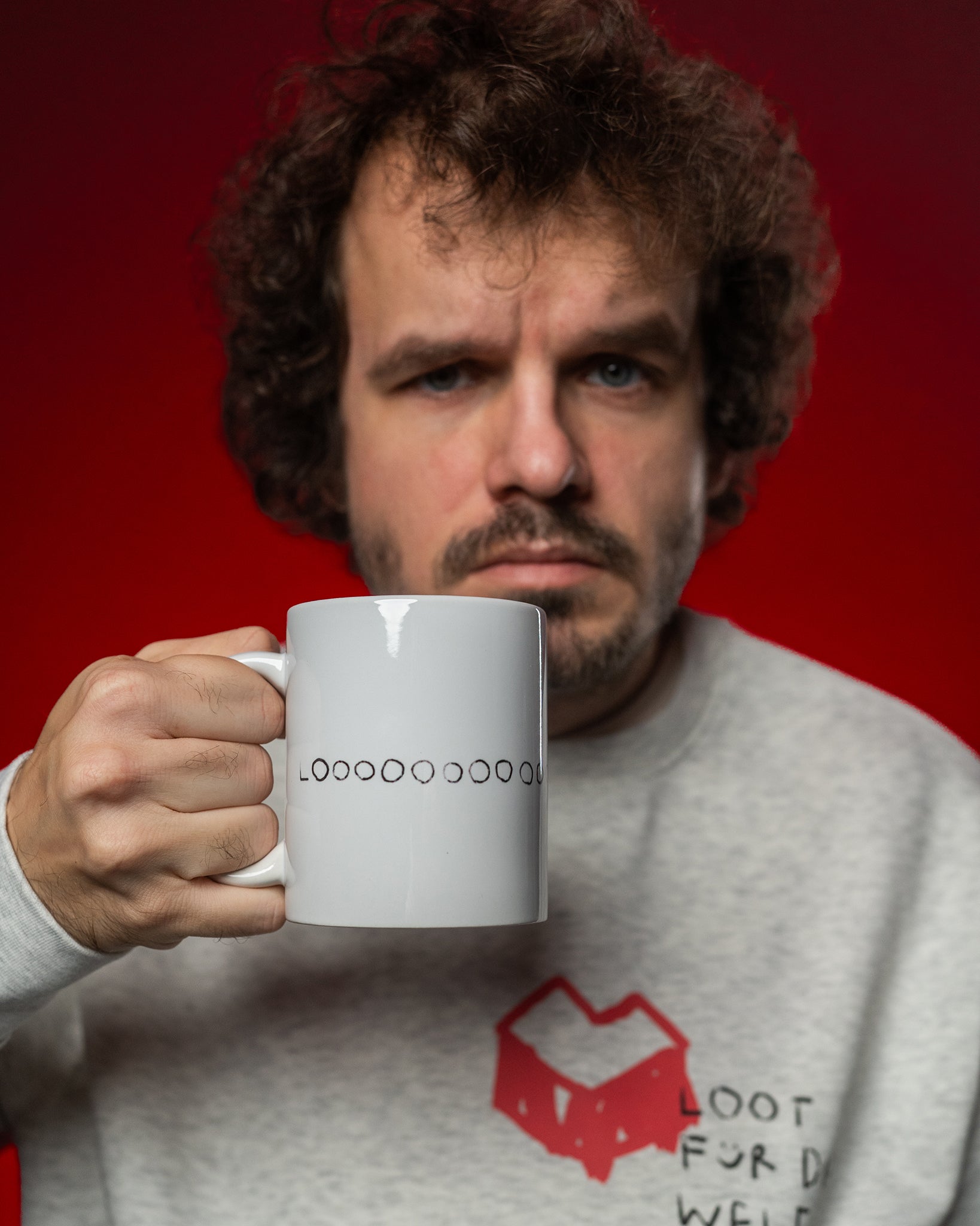 Eine Person hält eine weiße Keramiktasse mit dem schwarzen Schriftzug „LOOOOOOOOOOOT“ in der Hand. Die Tasse wird nach vorn präsentiert. Die Person trägt ein hellgraues Sweatshirt mit rotem, herzförmigem Logo und dem Text „LOOT FOR DA WELF“ auf der Brust. Der Hintergrund ist rot ausgeleuchtet. Der Fokus liegt auf der Tasse im Vordergrund.