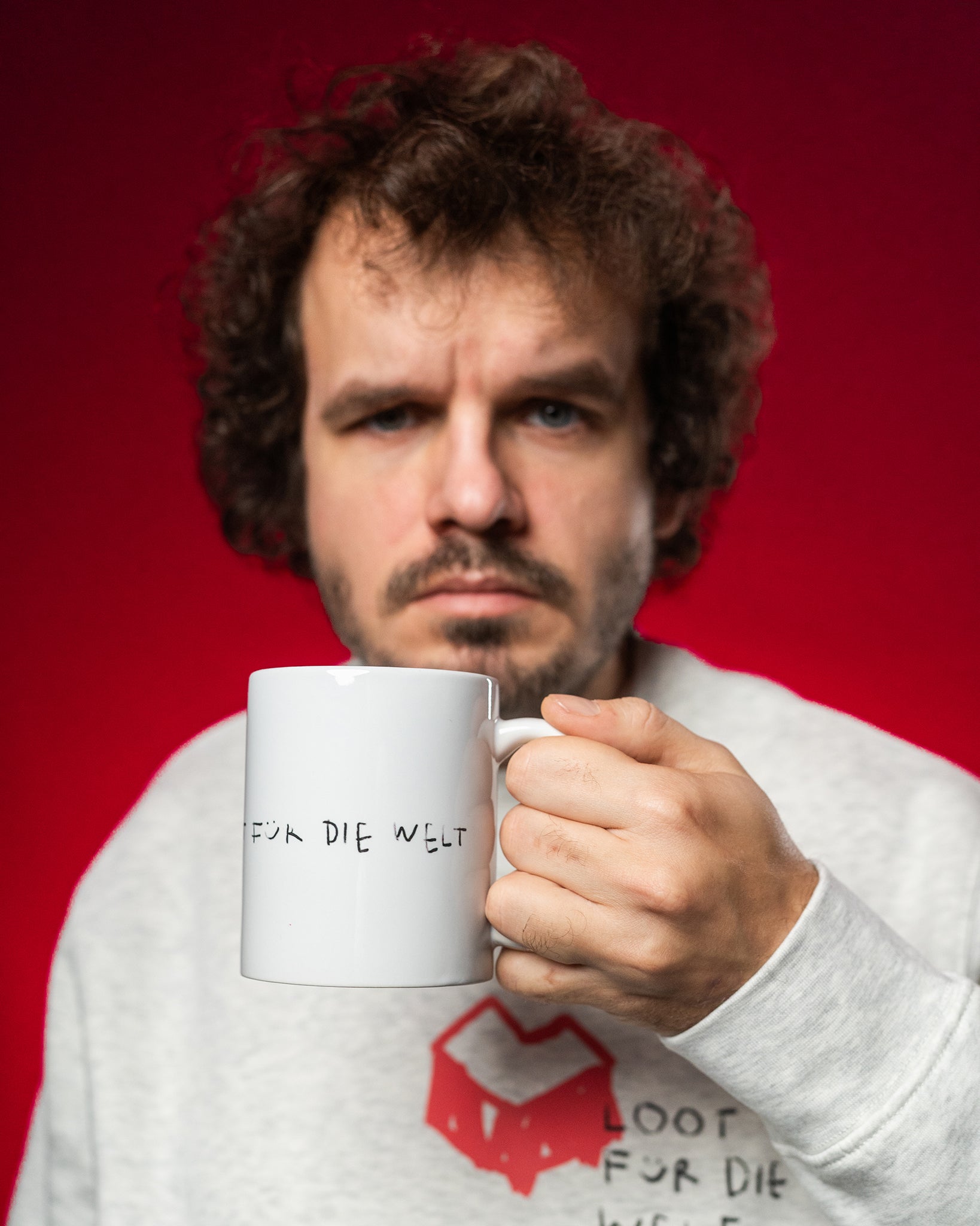 Eine Person hält eine weiße Keramiktasse mit schwarzer, handschriftlicher Aufschrift „FÜR DIE WELT“ nach vorn. Die Person trägt ein hellgraues Sweatshirt mit rotem, herzförmigem Logo und dem Text „LOOT FÜR DIE WELT“ auf der Brust. Der Hintergrund ist leuchtend rot, der Fokus liegt auf der Tasse im Vordergrund.