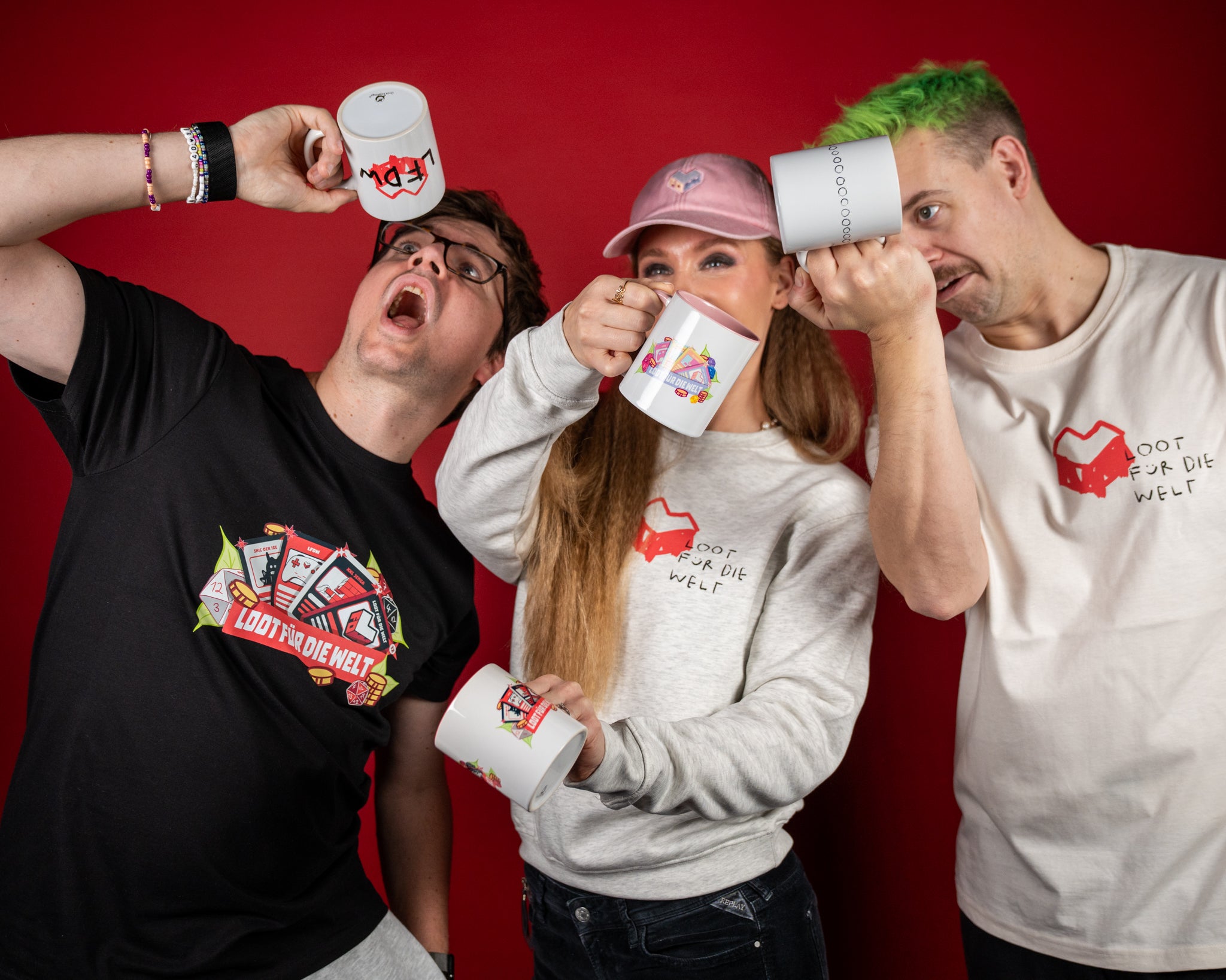 Drei Personen posieren fröhlich vor rotem Hintergrund und halten weiße „LOOT FÜR DIE WELT“-Tassen hoch. Eine Person trägt ein schwarzes T-Shirt mit farbigem Gaming-Motiv, eine hellgraues Sweatshirt mit rotem Herzlogo und eine beiges T-Shirt mit gleichem Logo. Die Szene zeigt spielerisch präsentierte Merch-Produkte der „LOOT FÜR DIE WELT“-Kollektion.
