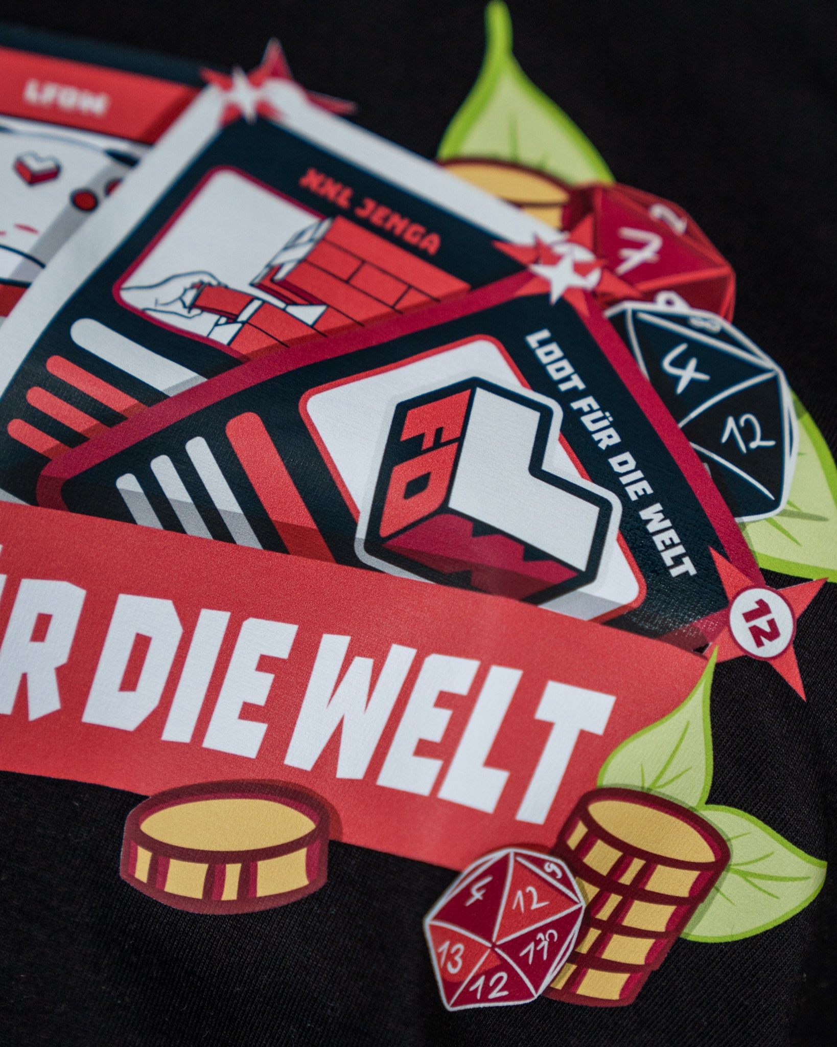 Detailaufnahme des Frontprints auf einem schwarzen T-Shirt. Das Motiv zeigt mehrere Spielkarten mit roten und schwarzen Elementen, auf denen Symbole wie ein Controller, Bauklötze und das „LFDW“-Logo zu sehen sind. Darunter verläuft ein rotes Banner mit der weißen Aufschrift „LOOT FÜR DIE WELT“. Ergänzt wird das Design durch goldene Münzen, bunte Würfel und grüne Blätter, die das verspielte Gaming-Thema betonen.
