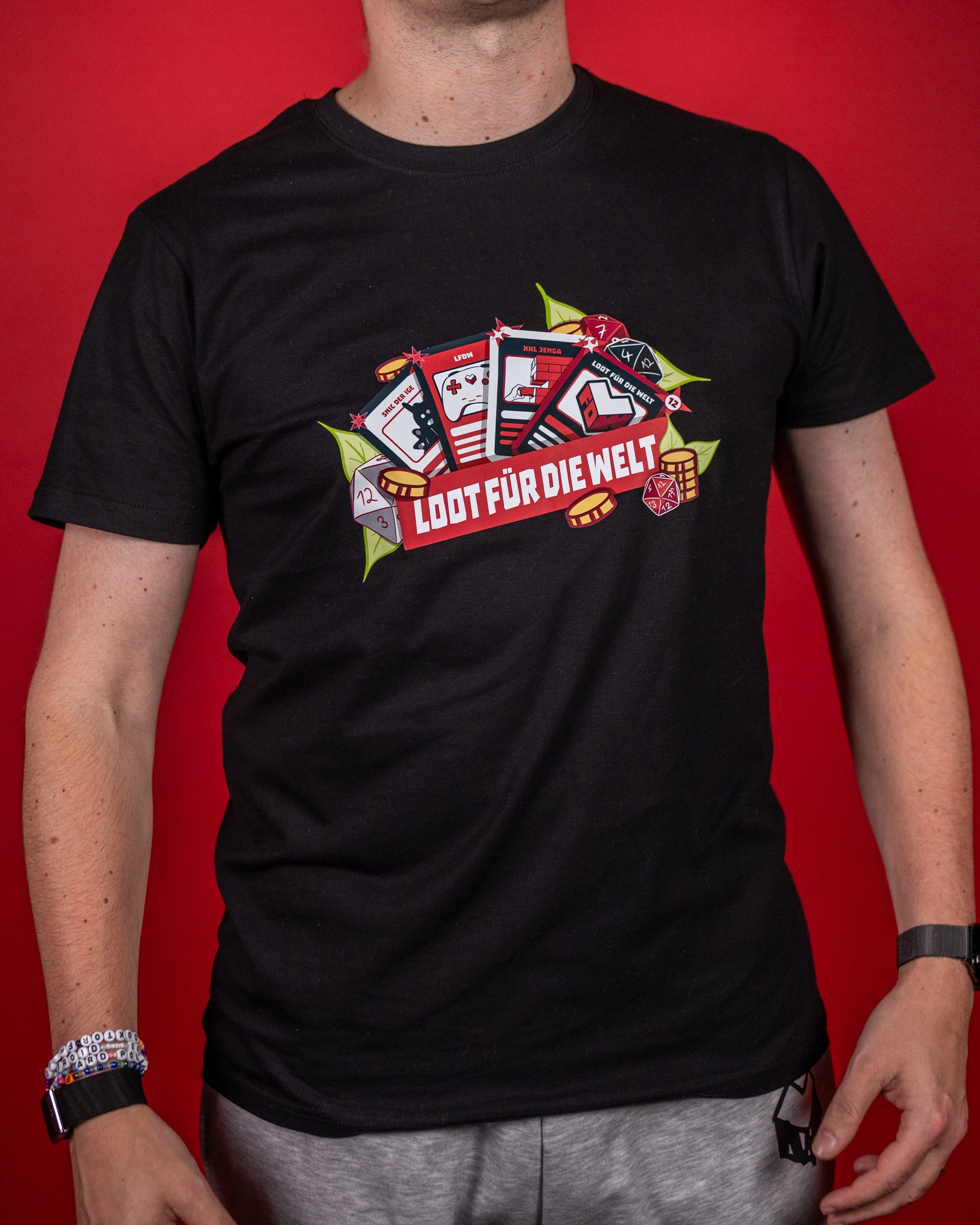 Schwarzes T-Shirt mit großem, farbigem Frontprint. Das Motiv zeigt mehrere Spielkarten mit Symbolen wie Controller, Würfel und Herz, ergänzt durch goldene Münzen und Blätter. In der Mitte steht der Schriftzug „LOOT FÜR DIE WELT“ auf einem roten Banner. Das Design kombiniert Gaming- und Charity-Themen in einem markanten, modernen Stil.