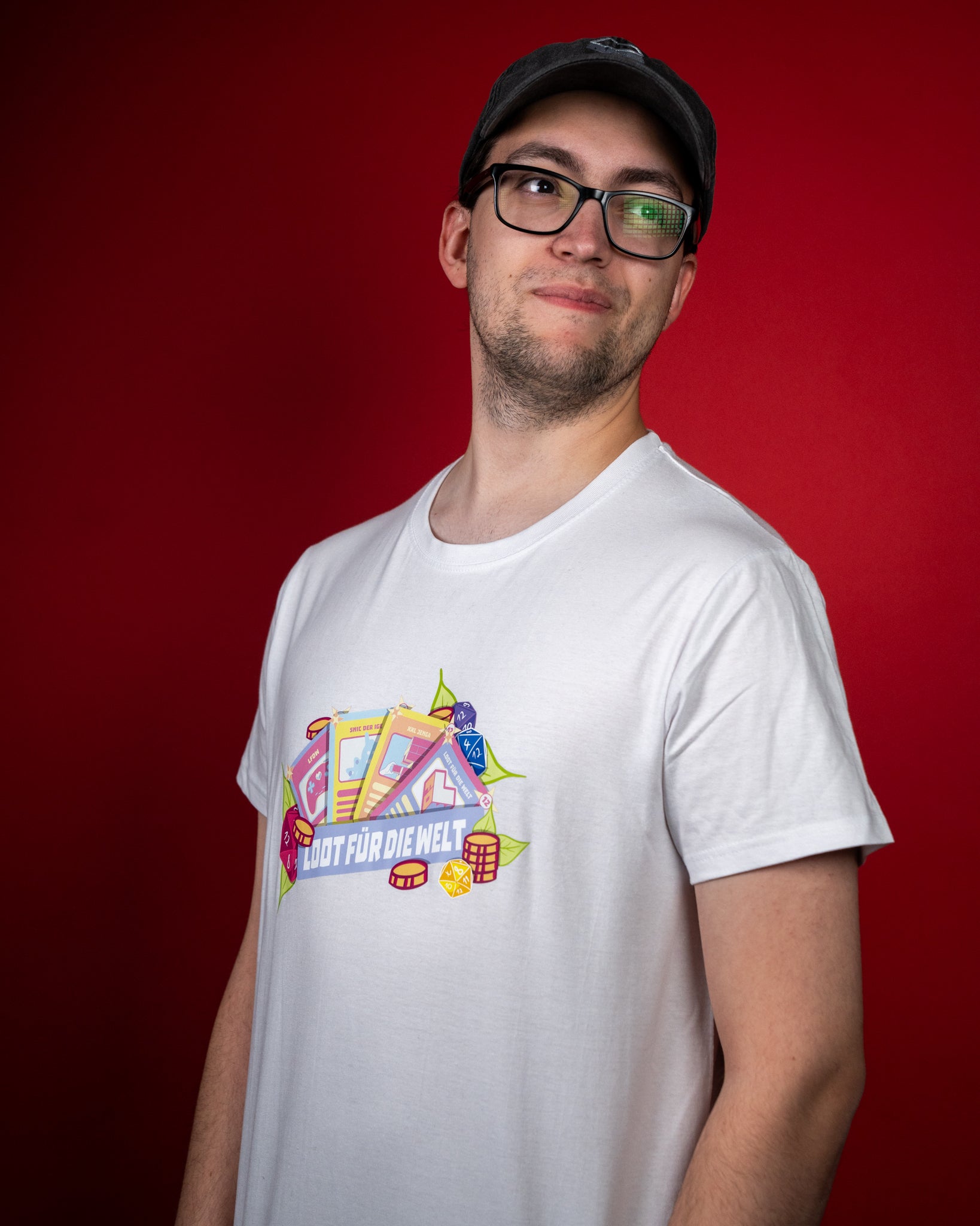 Weißes T-Shirt mit farbenfrohem Frontprint. Das Motiv zeigt stilisierte Karten, Würfel und goldene Münzen, die von grünen Blättern umrahmt werden. In der Mitte steht der Schriftzug „LOOT FÜR DIE WELT“ auf einem blauen Banner. Das Design kombiniert verspielte Gaming-Elemente mit einem frischen, illustrativen Look und vermittelt Freude, Teamgeist und Kreativität.
