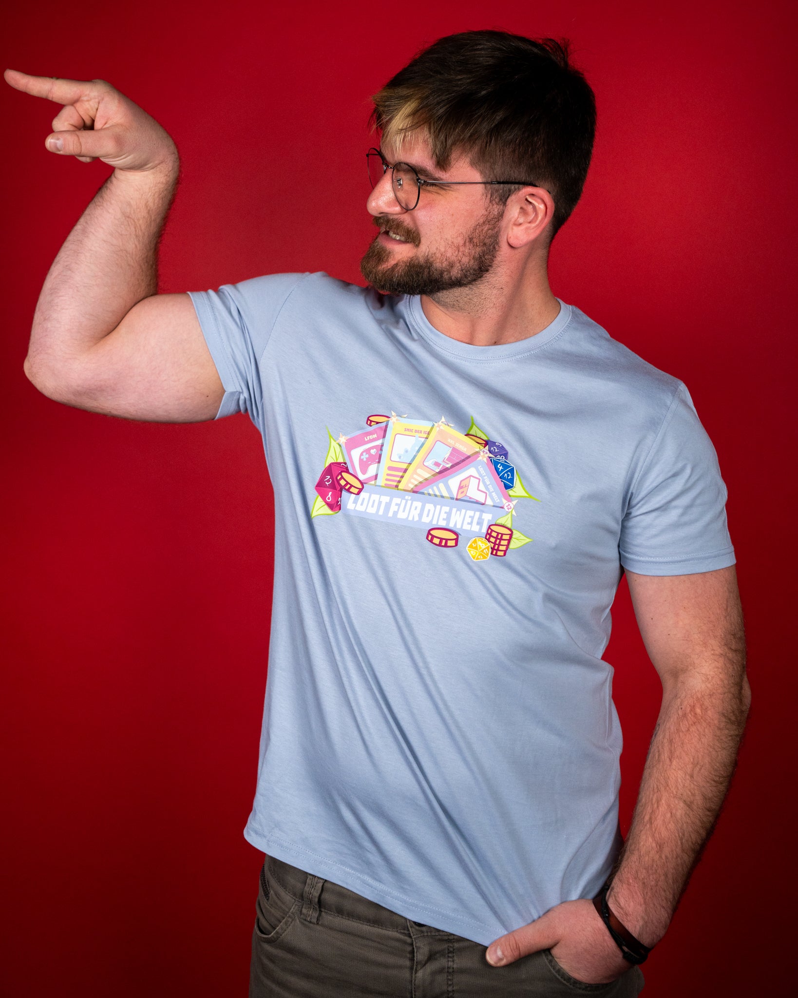 Hellblaues T-Shirt mit farbigem Frontprint im Stil einer Spielesammlung. Das Motiv zeigt mehrere bunte Karten mit Symbolen wie einem Gamecontroller, Bauklötzen und einer Spielfigur, umgeben von goldenen Münzen, Würfeln und grünen Blättern. Zentral darüber verläuft ein lilafarbenes Banner mit der weißen Aufschrift „LOOT FÜR DIE WELT“. Das Design wirkt verspielt und farbenfroh und steht in lebhaftem Kontrast zum hellen Stoff des Shirts.