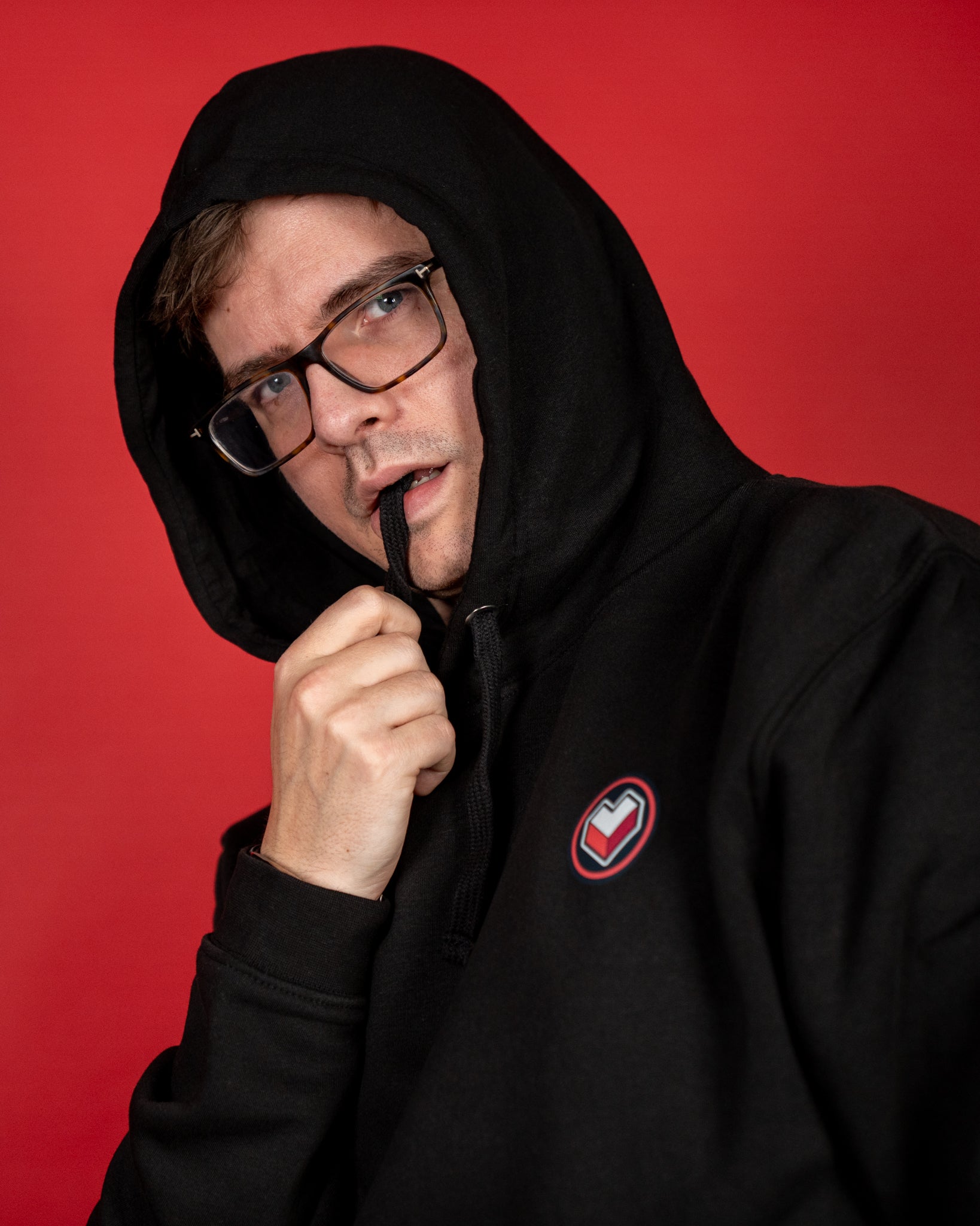 Nahaufnahme eines schwarzen Hoodies mit hochgezogener Kapuze. Auf der linken Brust ist ein rundes Logo mit stilisiertem Herzsymbol in Rot, Weiß und Grau zu sehen. Der Stoff wirkt weich und dicht, die Kapuze ist mit einem schwarzen Kordelzug ausgestattet. Der Hoodie vermittelt einen gemütlichen und modernen Look.