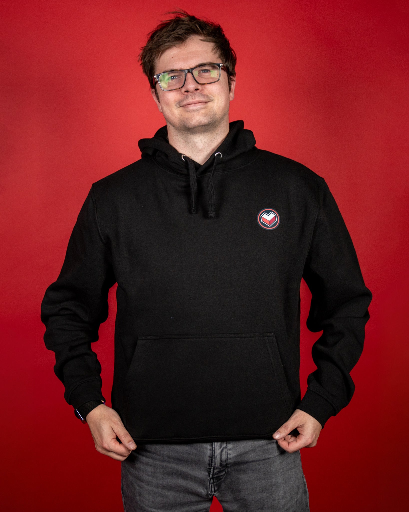 Schwarzer Hoodie mit kleinem, runden Brustlogo in Rot, Weiß und Grau. Das Motiv zeigt ein stilisiertes Herzsymbol im Kreis. Der Hoodie verfügt über eine große Kängurutasche, eine doppellagige Kapuze mit Kordelzug und elastische Bündchen an Ärmeln und Saum. Der Stoff wirkt weich und sorgt für einen bequemen Sitz.