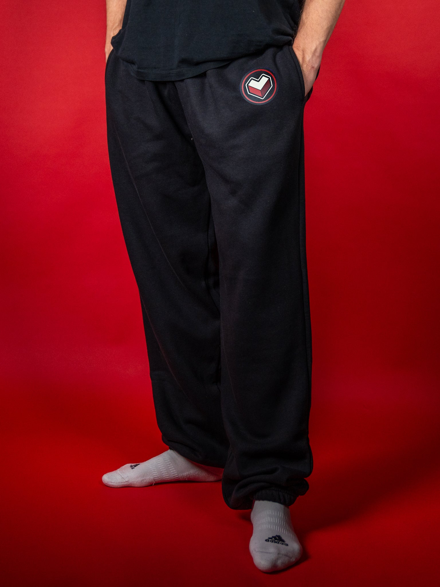 Schwarze Jogginghose aus weichem Stoff mit elastischem Bund und Bündchen an den Knöcheln. Auf der linken Seite befindet sich ein rundes Logo mit rotem Rand und stilisiertem Herzsymbol in Weiß und Grau. Die Hose hat eine lockere Passform und seitliche Eingrifftaschen – ideal für gemütliche Alltagslooks oder entspanntes Gaming.