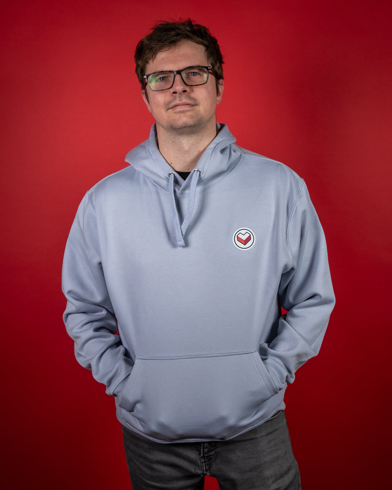 Hellblauer Hoodie aus weicher Baumwollmischung mit klassischer Kängurutasche und verstellbarer Kapuze. Auf der linken Brust befindet sich ein rundes Logo mit rotem Herzsymbol in geometrischer Form. Der Stoff wirkt glatt und hochwertig, die Passform locker und bequem – ideal für Freizeit und Alltag.