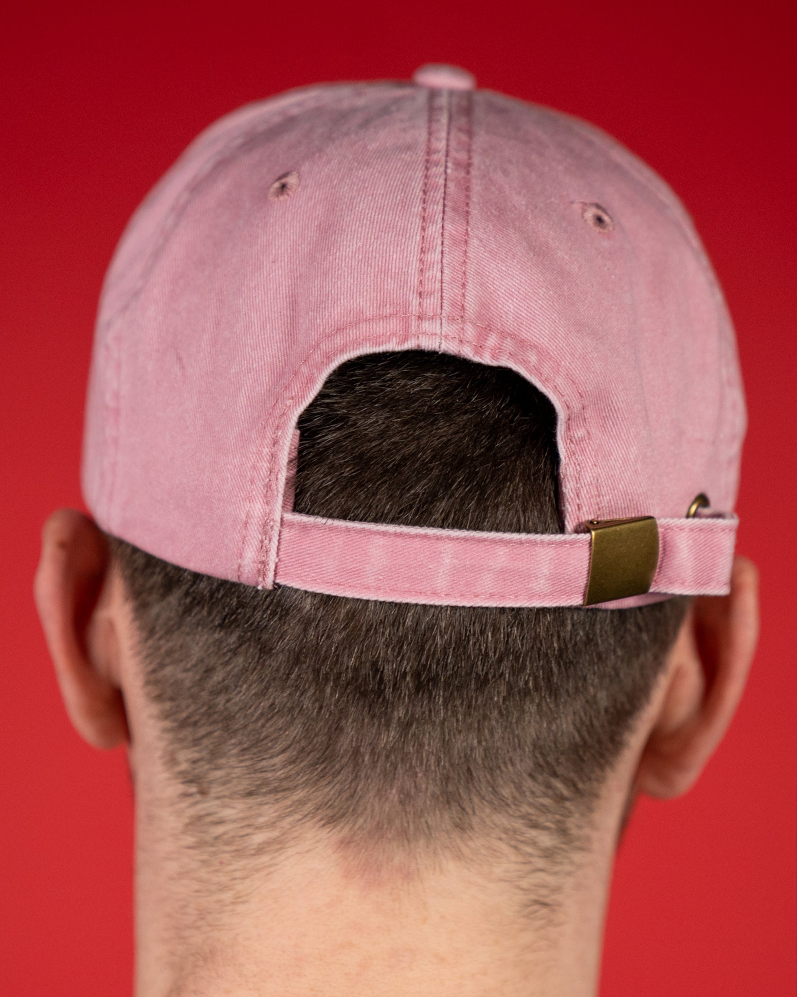 Rückansicht einer rosafarbenen Baumwoll-Cap mit verstellbarem Riemen aus Stoff und Metallschnalle in Messingoptik. Die Kappe zeigt sichtbare Ziernähte und Luftlöcher, die für eine klassische, luftige Passform sorgen.