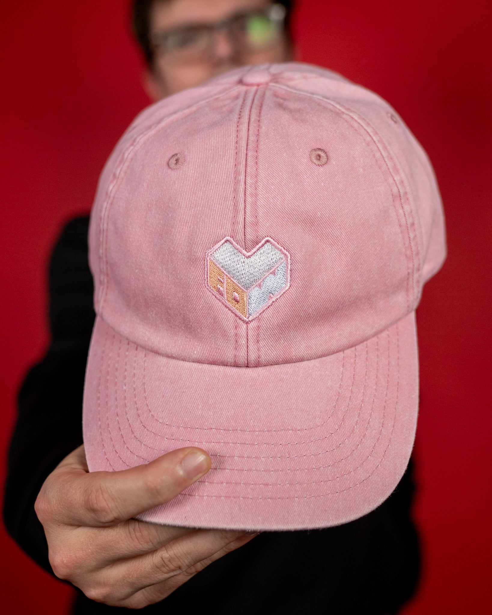 Nahaufnahme einer rosafarbenen Cap aus Baumwolle mit leicht gebogener Krempe. Auf der Vorderseite ist ein gesticktes Logo in Herzform zu sehen, das in Weiß und Pastellorange gehalten ist und die Buchstaben „LFDW“ zeigt. Die sichtbaren Nähte und Luftlöcher betonen den klassischen Baseballcap-Stil.