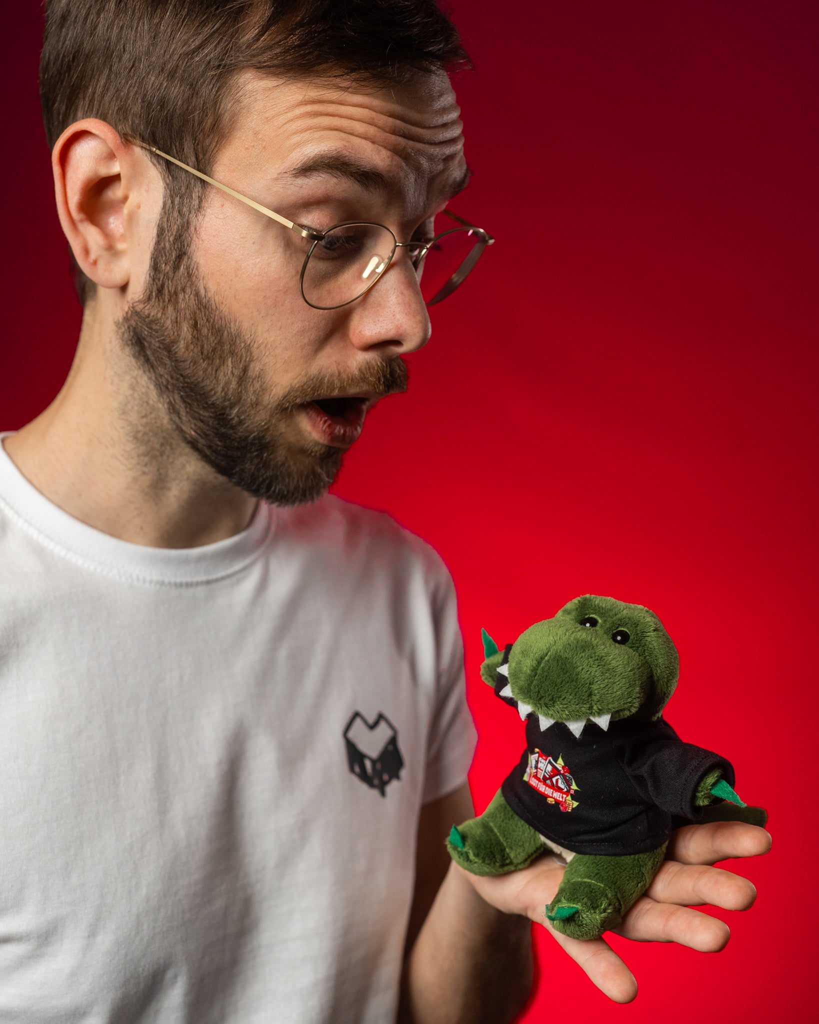 Kleiner grüner Plüsch-Dinosaurier mit flauschiger Oberfläche, weißen Stoffzähnen und gestickten Augen. Der Dino trägt ein schwarzes Mini-T-Shirt mit buntem „Loot für die Welt“-Logo, das Spielkarten, Münzen und Blätter zeigt. Er sitzt auf der Hand einer Person, die ihn mit überrascht-freundlichem Blick betrachtet. Der rote Hintergrund hebt den Dino und das farbenfrohe Shirtmotiv kontrastreich hervor. Das Kuscheltier wirkt hochwertig verarbeitet und detailreich gestaltet – ideal als Fanartikel oder Geschenk.