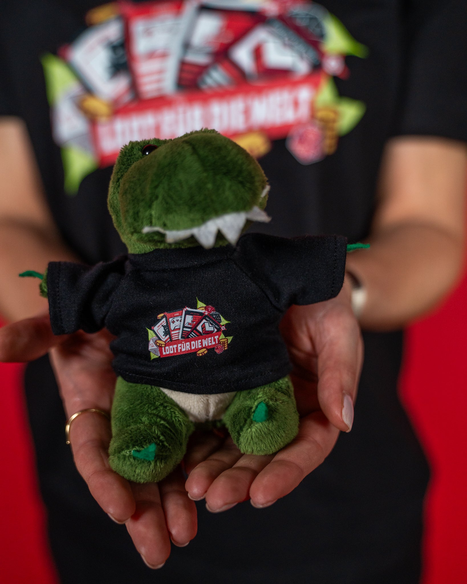 Kleiner grüner Plüsch-Dinosaurier mit weichem, flauschigem Stoff und weißen Stoffzähnen. Das Kuscheltier trägt ein schwarzes Mini-T-Shirt mit farbigem „Loot für die Welt“-Logo auf der Brust. Das Logo zeigt stilisierte Spielkarten, Münzen und Blätter in kräftigen Farben. Der Dino wird von zwei Händen nach vorne gehalten und wirkt freundlich und detailreich gestaltet. Ein liebevolles Sammlerstück oder Geschenk mit Bezug zur „Loot für die Welt“-Aktion.