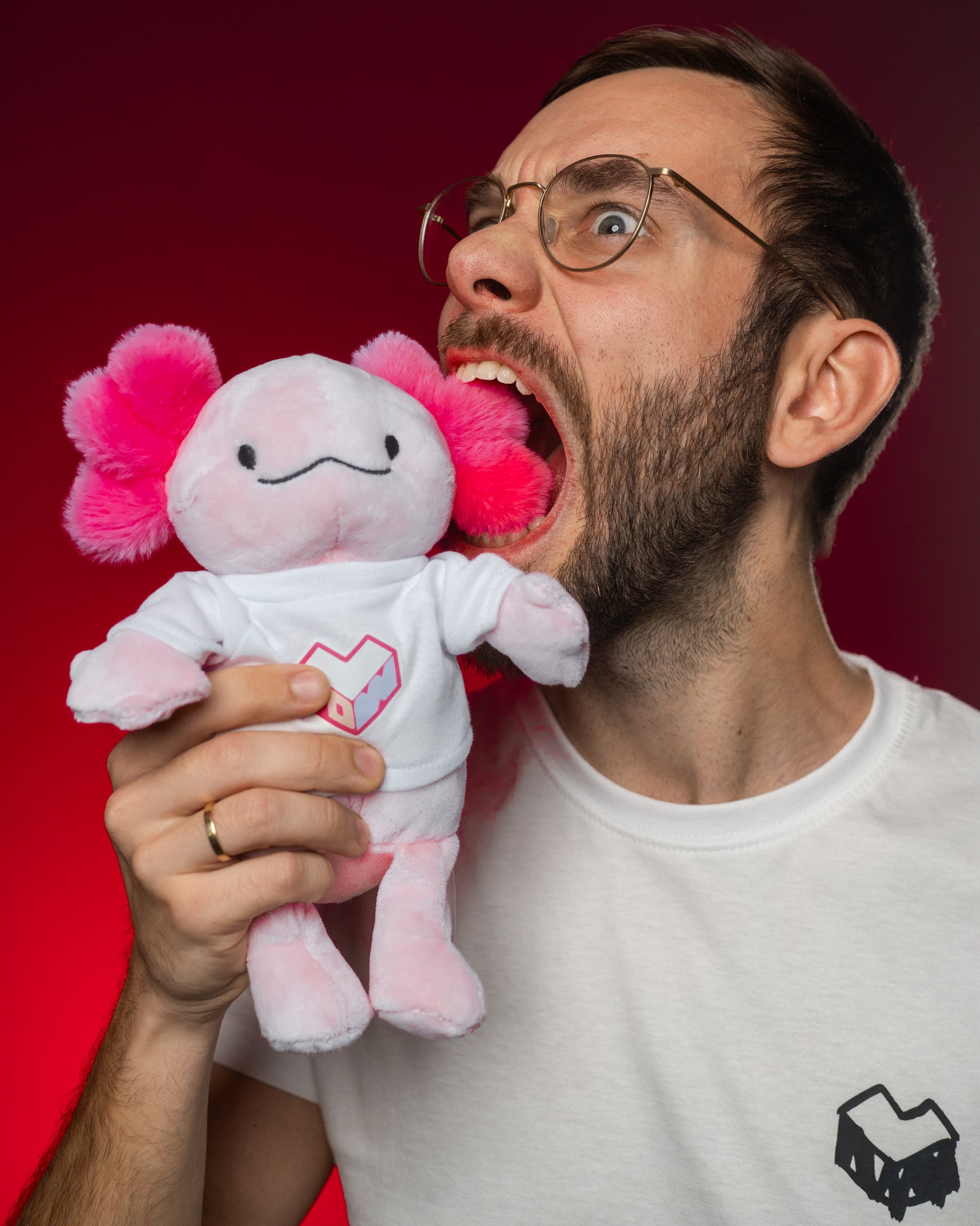 Rosa Plüsch-Axolotl mit pinken Kiemenbüscheln und weißem Shirt mit rosa-violettem Herzlogo „LFDW“. Das Stofftier wird humorvoll in die Kamera gehalten, der rote Hintergrund hebt die leuchtenden Farben hervor. Der Fokus liegt auf dem detailreich gestalteten Fan-Plüschie.