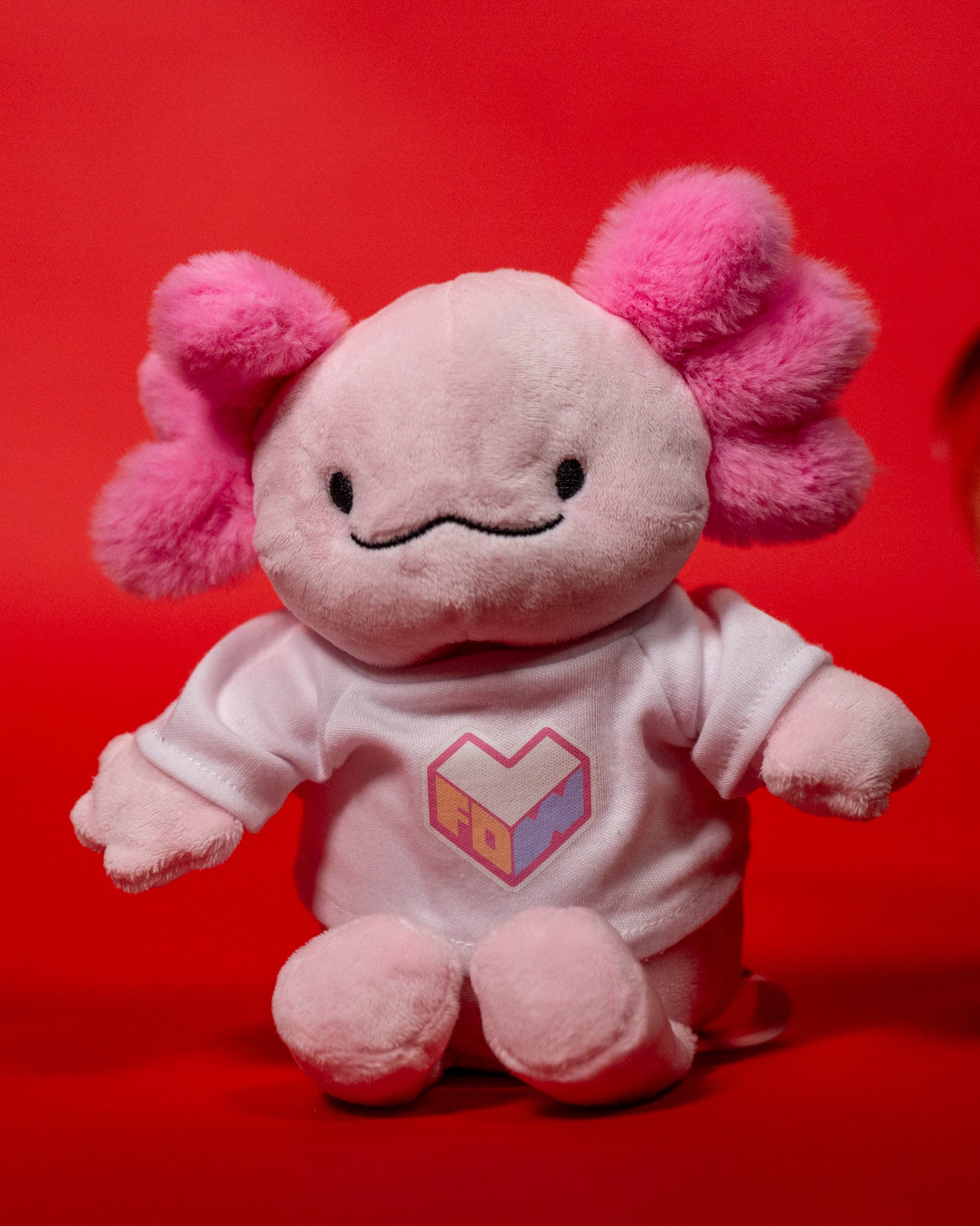 Rosa Plüsch-Axolotl mit flauschigen pinken Kiemenbüscheln und freundlichem Gesicht. Das Stofftier trägt ein weißes Shirt mit rosa-violettem Herzlogo „LFDW“. Der rote Hintergrund betont die weiche Struktur und den verspielten Charakter des Fanartikels.