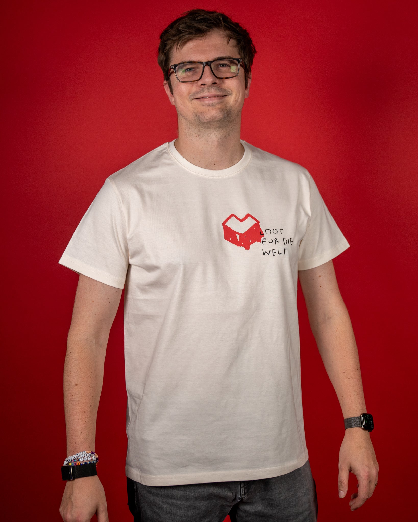 Cremefarbenes T-Shirt mit kleinem Frontprint auf der linken Brustseite. Das Motiv zeigt das rote „Loot für die Welt“-Herzlogo, daneben steht in schwarzer, handschriftlich wirkender Schrift „LOOT FÜR DIE WELT“. Das Shirt hat einen klassischen Rundhalsausschnitt und kurze Ärmel. Es wird locker getragen, die Person steht aufrecht und lächelt leicht, wodurch der Print gut zur Geltung kommt. Der rote Hintergrund hebt das cremefarbene Shirt und das Motiv kontrastreich hervor.