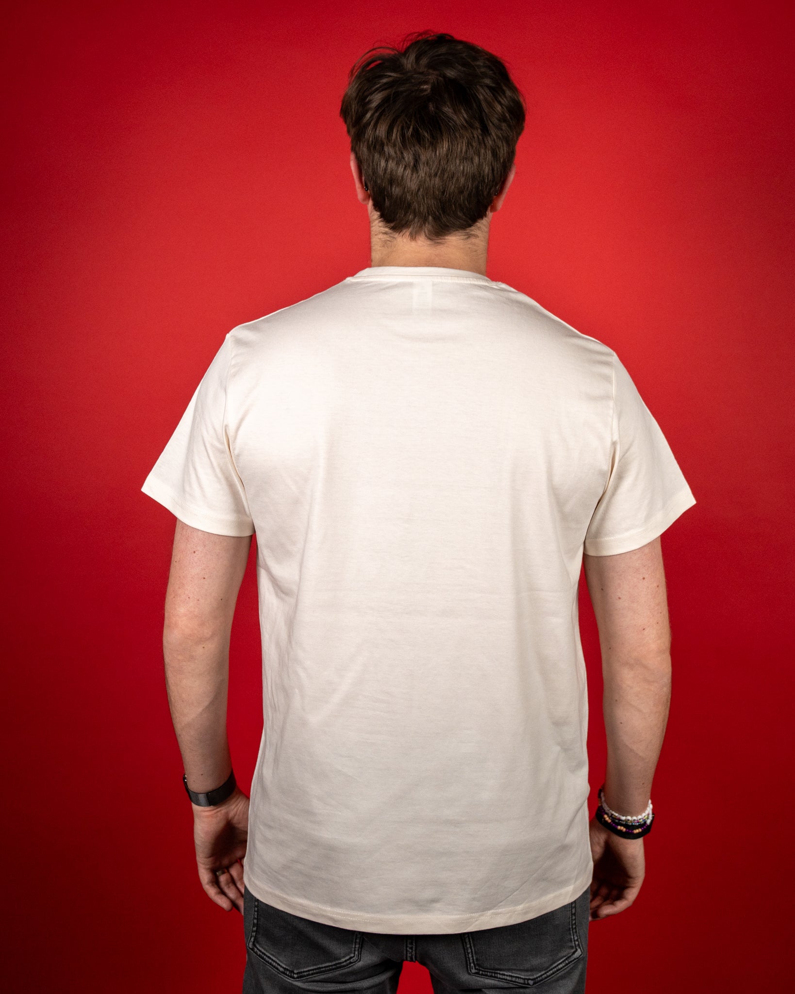 Rückansicht eines cremefarbenen T-Shirts ohne Aufdruck. Das Shirt hat einen klassischen Rundhalsausschnitt und kurze Ärmel. Der Stoff fällt glatt und gerade über Rücken und Schultern. Die Person steht locker mit herabhängenden Armen vor rotem Hintergrund, wodurch die schlichte Rückseite und die neutrale Farbgebung des T-Shirts klar zur Geltung kommen.