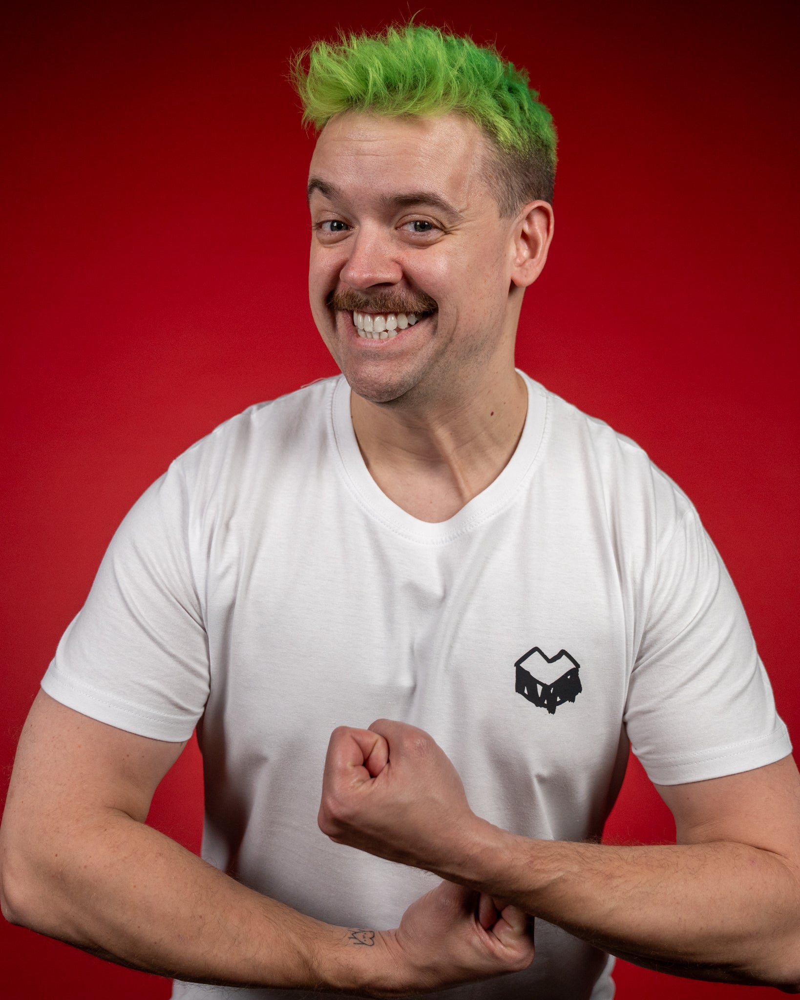 Weißes T-Shirt mit kleinem schwarzen Logo auf der linken Brustseite. Das Motiv zeigt ein kantiges Herzsymbol in einer würfelförmigen Box – das typische „Loot für die Welt“-Logo. Das T-Shirt hat kurze Ärmel, einen runden Halsausschnitt und eine gerade Passform. Der Stoff wirkt glatt und fein gewebt. Die Person steht vor rotem Hintergrund, lächelt breit und spannt dabei spielerisch die Armmuskeln an, wodurch das Shirt locker und natürlich präsentiert wird.