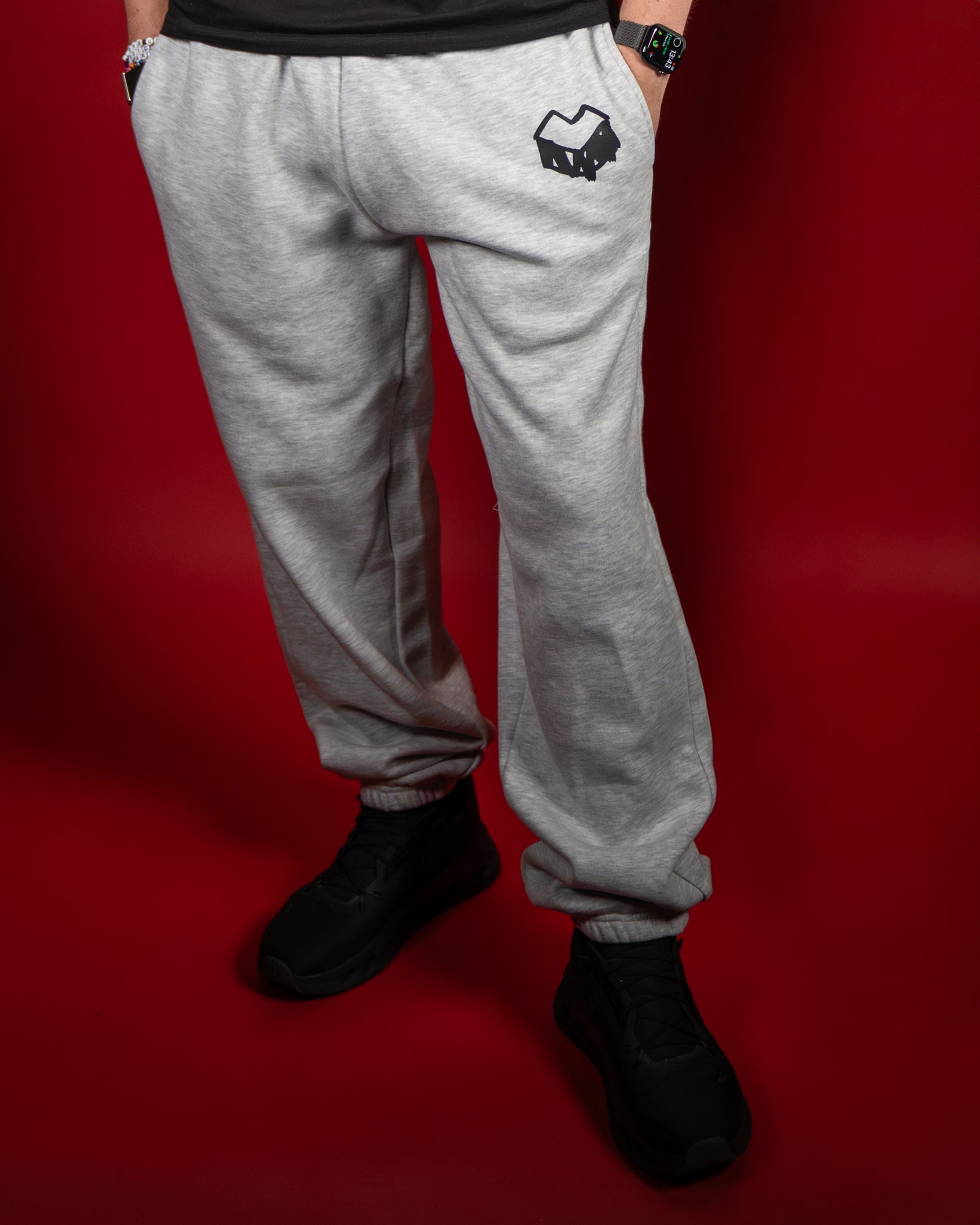 Graue Jogginghose aus weichem Sweatstoff mit elastischem Bund und Bündchen an den Knöcheln. Auf der linken Seite befindet sich ein schwarzes Logo in Form eines stilisierten Herzens. Die Hose fällt locker und sorgt für eine bequeme, lässige Passform – ideal für Freizeit, Sport oder gemütliche Tage zuhause.