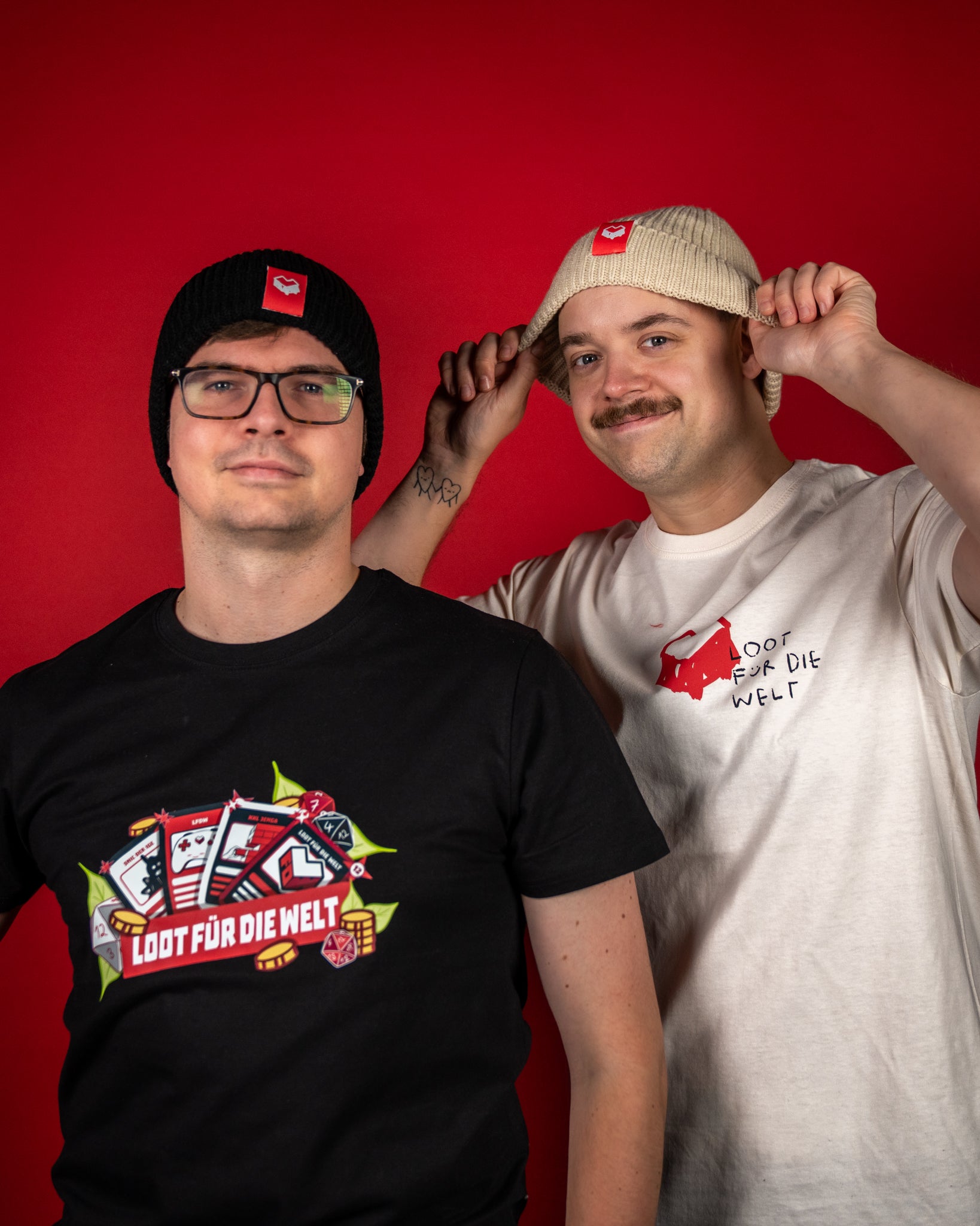 Zwei Rippstrick-Beanies in Schwarz und Beige, beide mit breiter Umschlagkante und rotem Label auf der Vorderseite. Auf dem Label ist das weiße „Loot für die Welt“-Logo abgebildet. Die schwarze Mütze wirkt klassisch und kontrastreich, während die beige Variante einen warmen, natürlichen Ton bietet. Beide Modelle bestehen aus weichem, elastischem Strickmaterial, das angenehm sitzt und zuverlässig warmhält – perfekt für kalte Tage und kombinierbar mit Streetwear- oder Casual-Looks.
