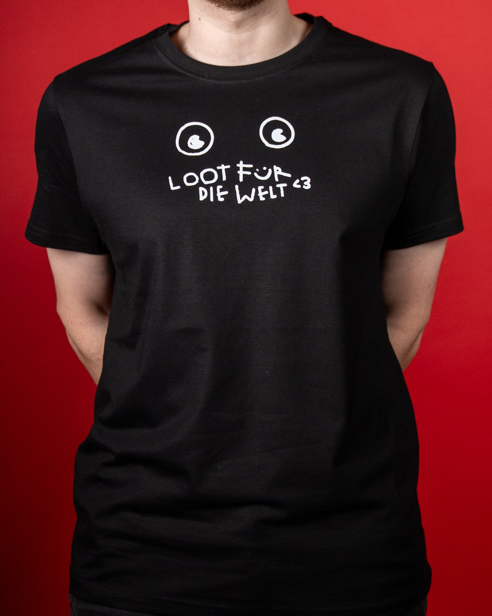 Schwarzes T-Shirt mit weißem Frontprint im handschriftlichen Stil. Das Motiv zeigt zwei stilisierte Augen oberhalb der Aufschrift „LOOT FÜR DIE WELT <3“, wobei das „Ü“ als lächelndes Gesicht gestaltet ist. Der Druck wirkt verspielt und erinnert an eine einfache, gezeichnete Comicfigur. Das Shirt hat einen klassischen Rundhalsausschnitt und kurze Ärmel, der Stoff fällt glatt über die Körperform. Der rote Hintergrund hebt den weißen Druck kontrastreich hervor.