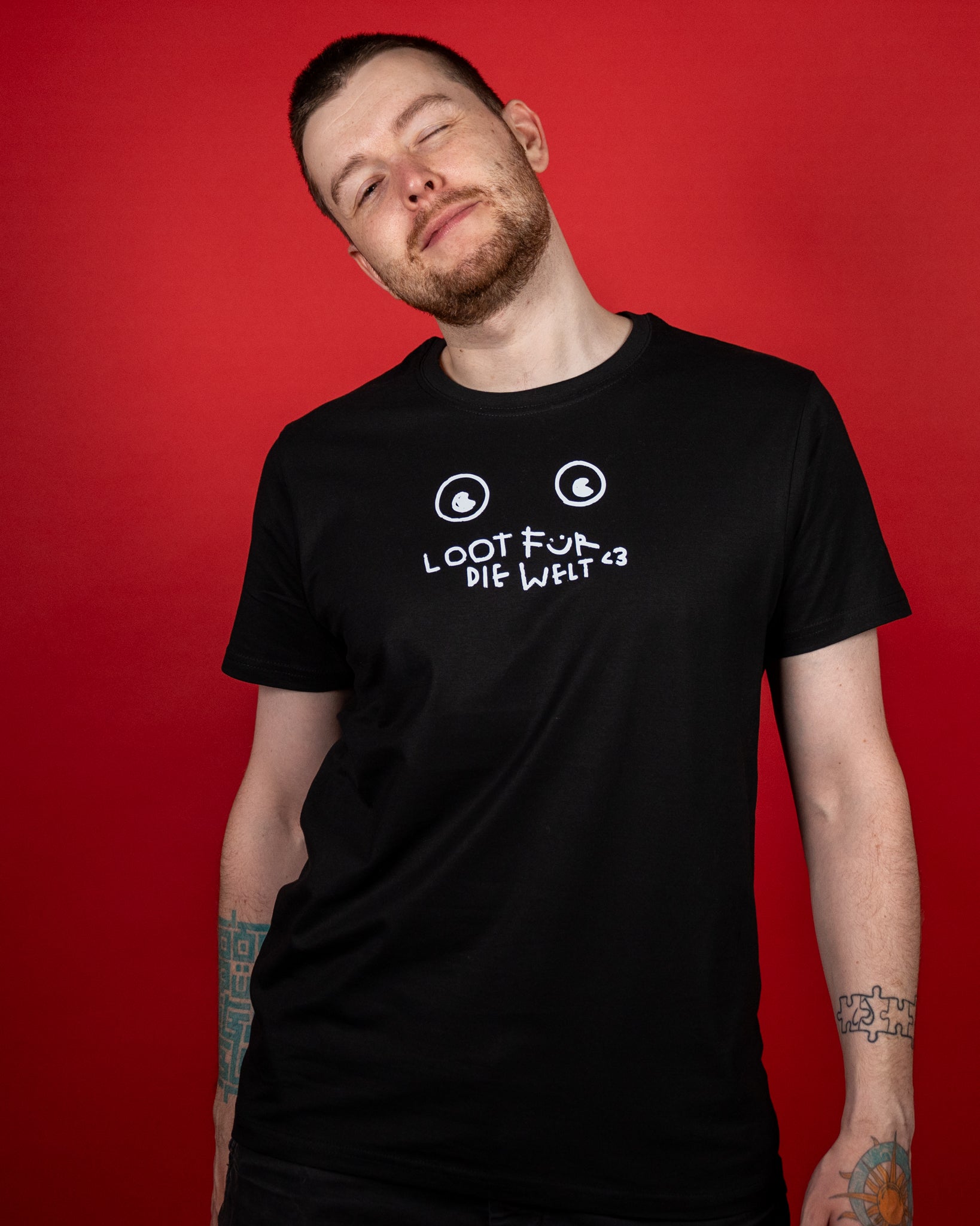 Schwarzes T-Shirt mit weißem Frontprint im handgezeichneten Stil. Der Aufdruck zeigt zwei runde Augen über der verspielten Aufschrift „LOOT FÜR DIE WELT <3“, wobei das „Ü“ als lächelndes Gesicht gestaltet ist. Das Shirt besitzt einen klassischen Rundhalsausschnitt und kurze Ärmel. Es wird leger getragen, während der Träger den Kopf leicht zur Seite neigt – so kommt der Druck auf der Brust gut zur Geltung. Der rote Hintergrund hebt das schwarze Shirt und den weißen Druck kontrastreich hervor.